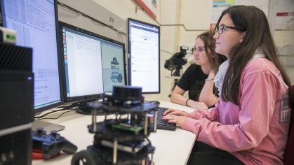 Two female college students sitting at desk looking at a computer. Girl in foreground with long dark hair and glasses is wearing a pink shirt. Girl in the background has brown hair with a black shirt. A robotic device is on the table.