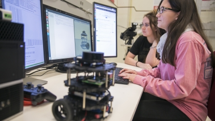 Two female college students sitting at desk looking at a computer. Girl in foreground with long dark hair and glasses is wearing a pink shirt. Girl in the background has brown hair with a black shirt. A robotic device is on the table.