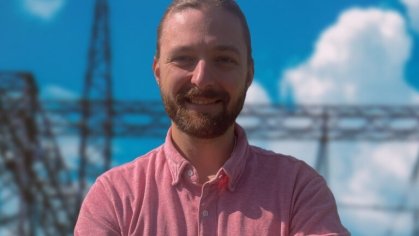 head shot of male with arms folded in front of a power grid with short blonde hair and moustache and beard, wearing a salmon colored button down shirt 