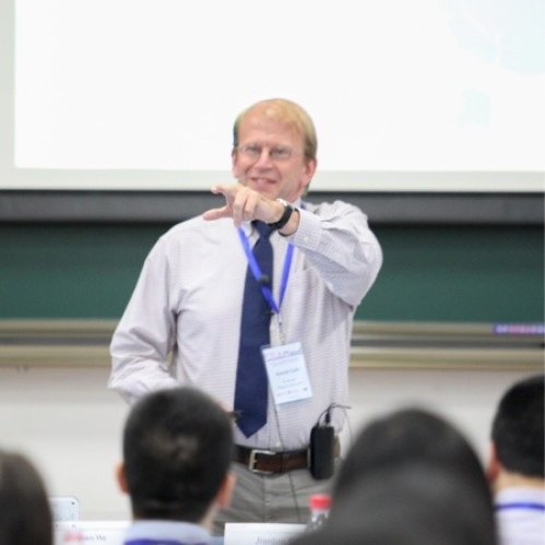 male wearing glasses, button down shirt, and blue tie lecturing to students and pointing