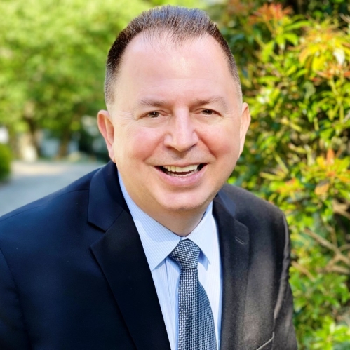 white male wearing a dark blue suit, light blue shirt, and patterned tie