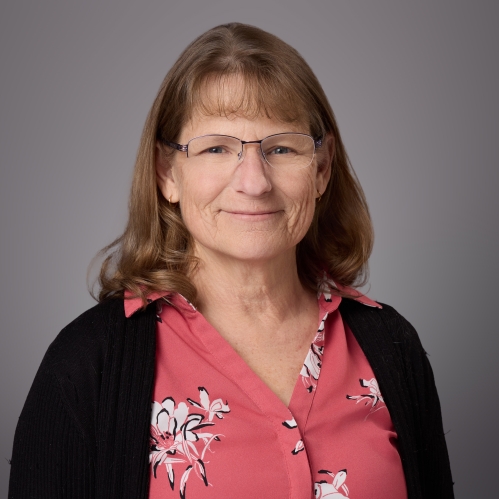 Headshot of female with light brown shoulder length hair, bangs, and eyeglasses wearing a red patterned blouse and a black sweater.