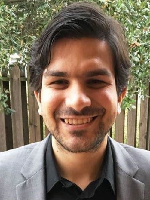 Smiling bearded man with dark brown hair wearing a grey suit jacket and black shirt