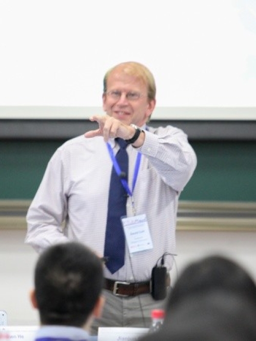 male wearing glasses, button down shirt, and blue tie lecturing to students and pointing