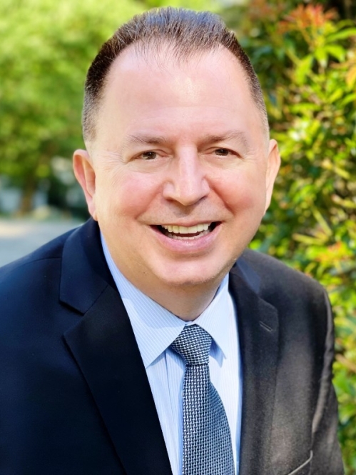 white male wearing a dark blue suit, light blue shirt, and patterned tie