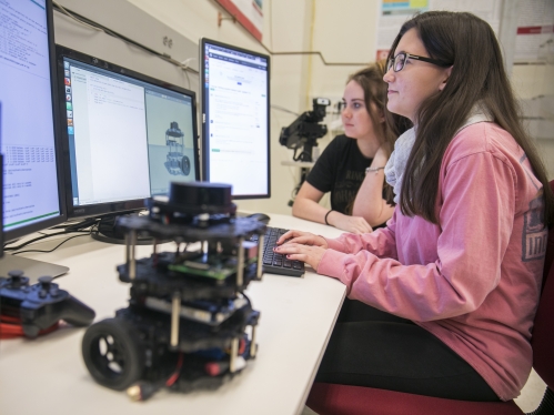 Two female college students sitting at desk looking at a computer. Girl in foreground with long dark hair and glasses is wearing a pink shirt. Girl in the background has brown hair with a black shirt. A robotic device is on the table.