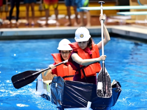 two women wearing white hardhats racing in a pool in a cardboard canoe 