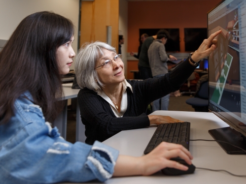An older woman professor with glasses and gray hair points to a computer screen as college student with black hair and a blue shirt controls the computer mouse and looks at the screen.