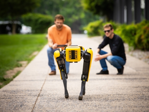 Outdoor photo of two men kneeling in the background behind a yellow robotic dog.