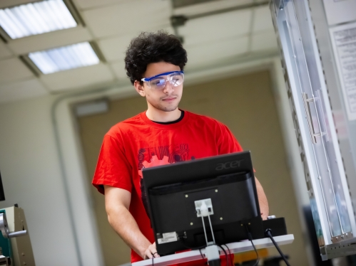 Male student with black curly hair poses behind a laptop. He's wearing a red T-shirt and protective eyewear.