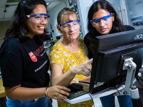 Two female students wearing black T-shirts standing on either side of a female faculty member wearing a yellow shirt, all wearing protective glasses looking at a computer monitor in a lab.