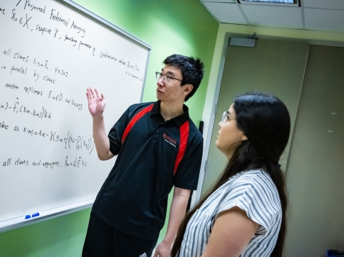Two graduate students stand looking at a whiteboard covered with mathematical formulas.