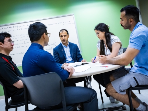 Professor wearing a dark blue suit and light blue open collared shirt, talks with four students around a round table. The students include on female, sitting to the professors left with long black hair and a white shirt, and three males.