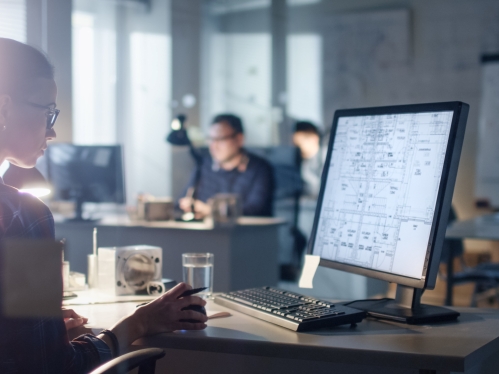 Young woman looks at computer screen in an office with others workers in the backgrounnd.