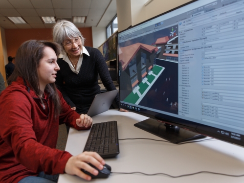 female professor with gray hair and eyeglasses looking at a computer screen, instructing a female student with long brown hair