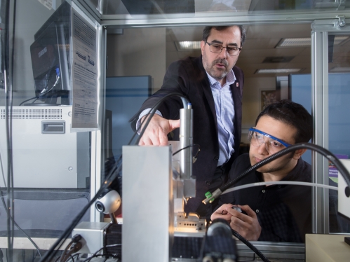 male professor with eyeglasses and beard working with machinery instructing a male student with dark hair wearing safety glasses