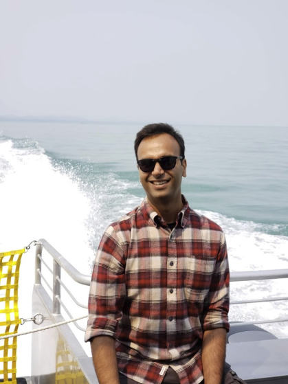 Man in a plaid shirt, dark hair and sunglasses, sits at the rear of a moving boat with an ocean wake behind.