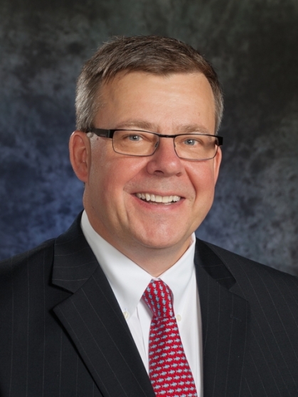 Head shot of white male wearing a dark suit with a white shirt and red tie.