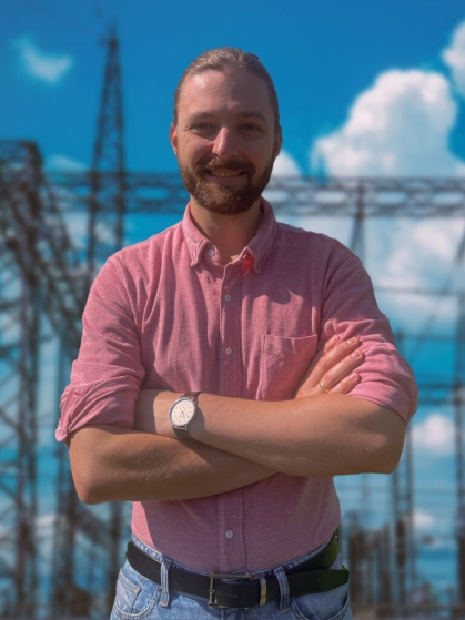head shot of male with arms folded in front of a power grid with short blonde hair and moustache and beard, wearing a salmon colored button down shirt